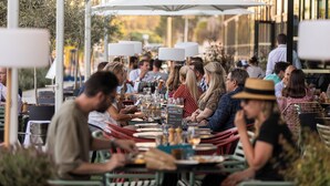 An outdoor dining area with tables and chairs, umbrellas, and a crowd of people.