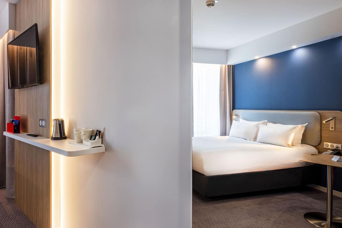 Ein modernes Hotelzimmer mit einem Bett, einem Nachttisch, einem an der Wand montierten Flachbildfernseher und einer kleinen Ablage mit Wasserkocher und Tassen.