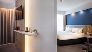 A modern hotel room with a bed, a bedside table, a flat-screen TV mounted on the wall, and a small shelf with a kettle and cups. - Holiday Inn Express Nice Grand Arenas by IHG (Nice)