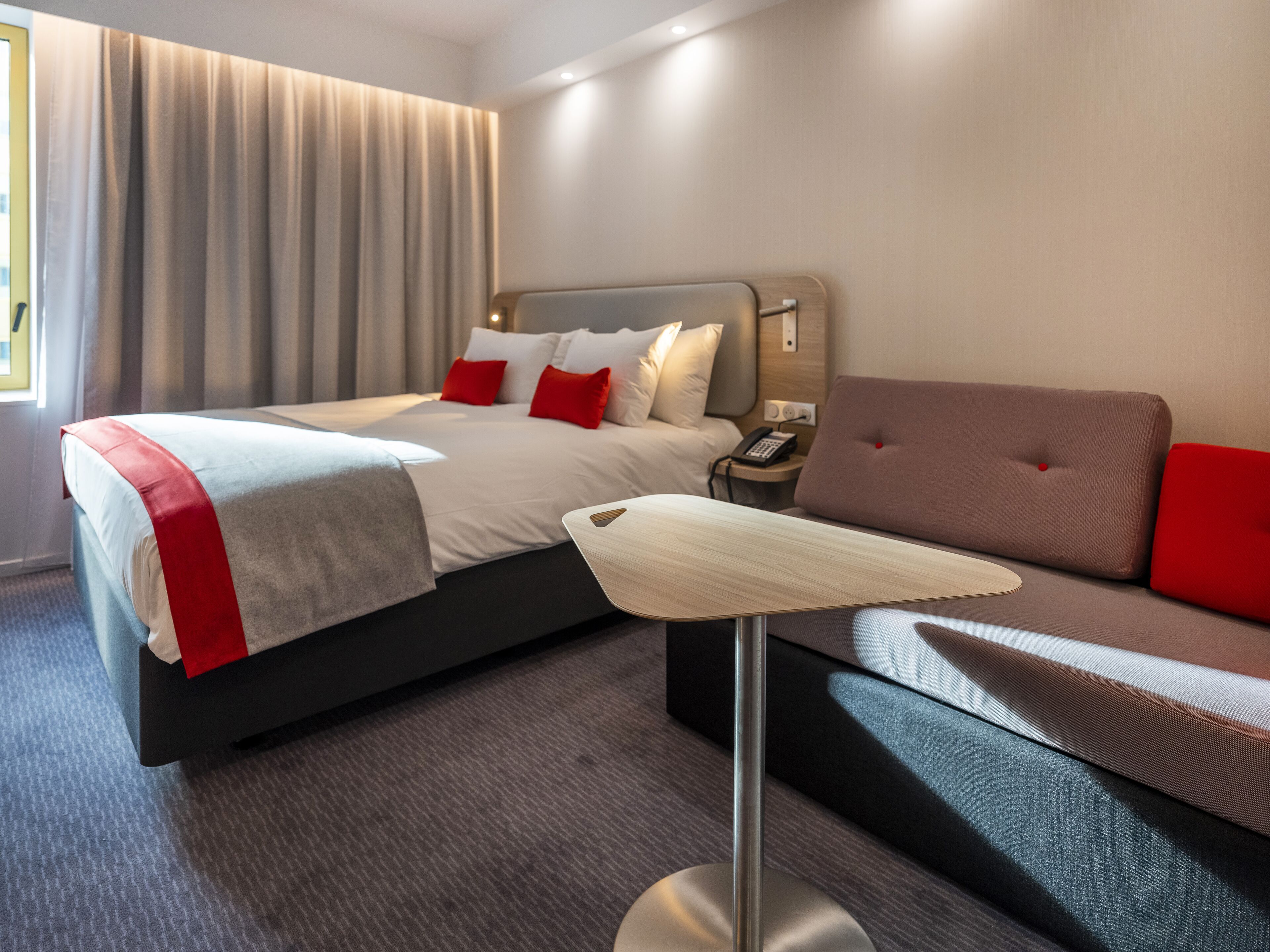 Ein modernes Hotelzimmer mit einem ordentlich bezogenen Bett, einem Sofa, einem Nachttisch mit Telefon und einer Lampe.