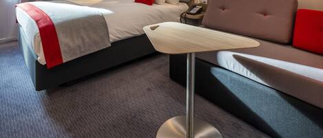 Ein modernes Hotelzimmer mit einem ordentlich bezogenen Bett, einem Sofa, einem Nachttisch mit Telefon und einer Lampe.