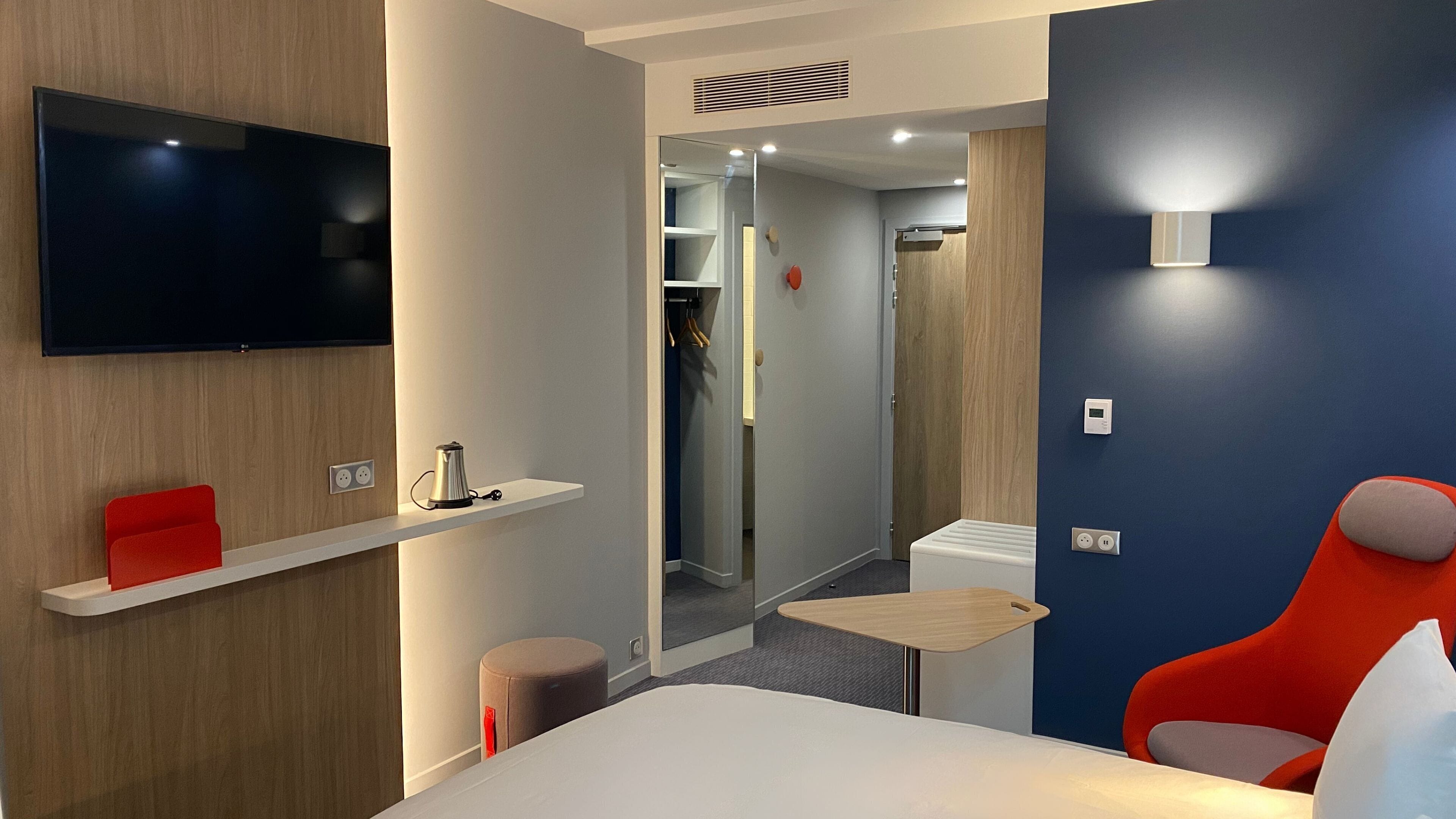 A modern hotel room with a bed, a TV mounted on the wall, a small table, a red chair, and a shelf with a kettle.