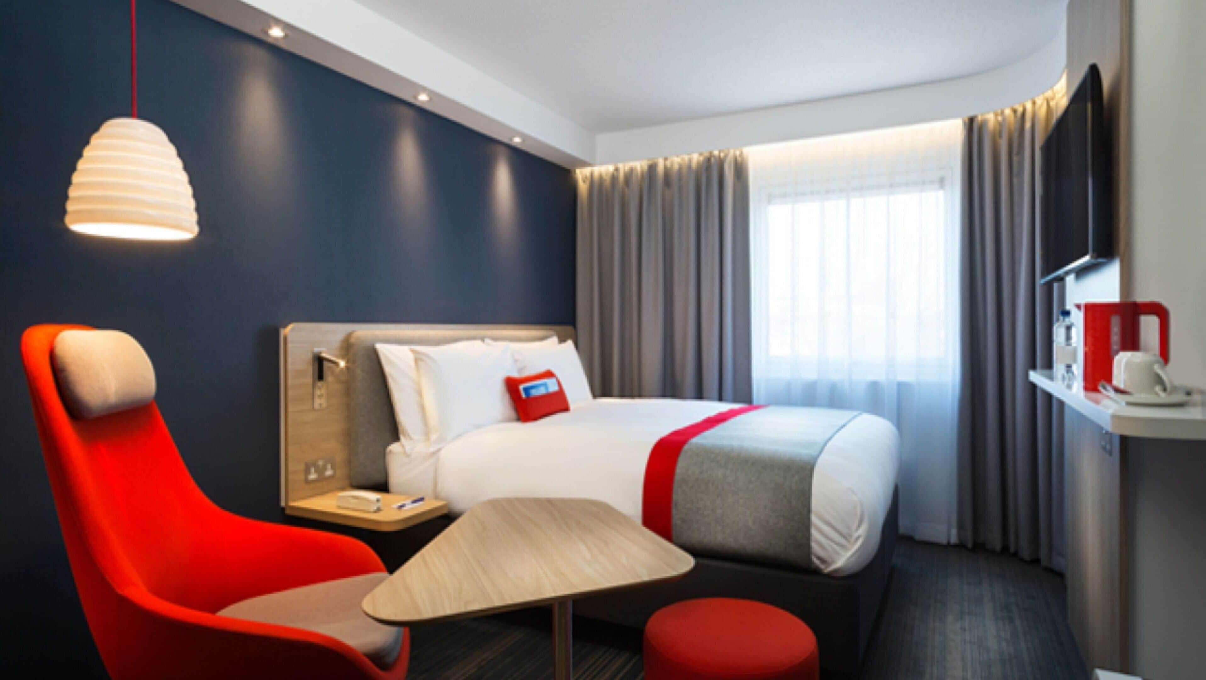 A hotel room with a bed, a desk, a chair, a table, and a hanging light fixture.