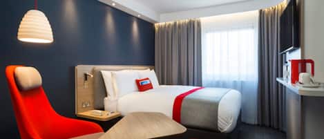 A hotel room with a bed, a desk, a chair, a table, and a hanging light fixture.