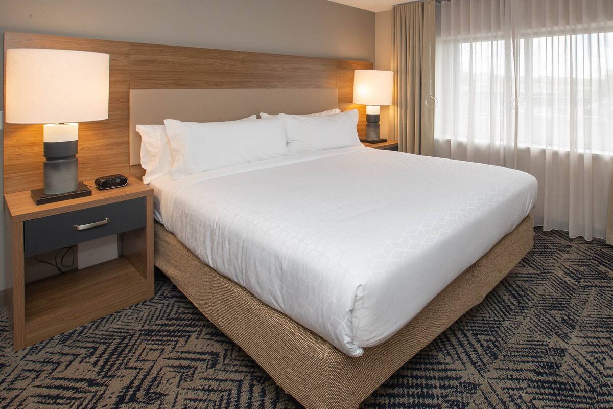 Habitación de hotel con una cama grande, dos mesitas de noche con lámparas, un televisor y una ventana con cortinas.
