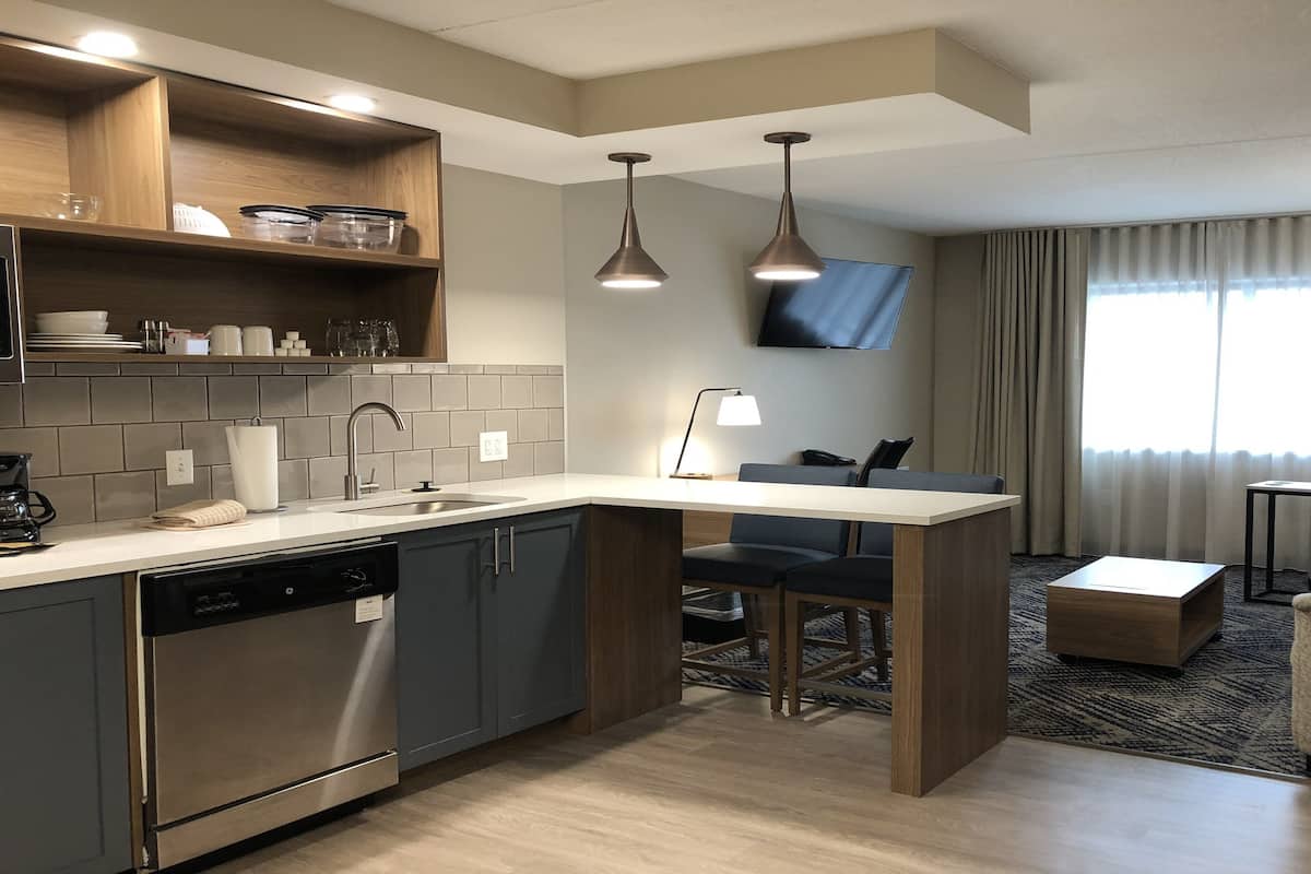 Una habitación de hotel moderna con cocina, zona de estar con sofá y sillones, televisión de pantalla plana montada en la pared y un gran ventanal con cortinas.