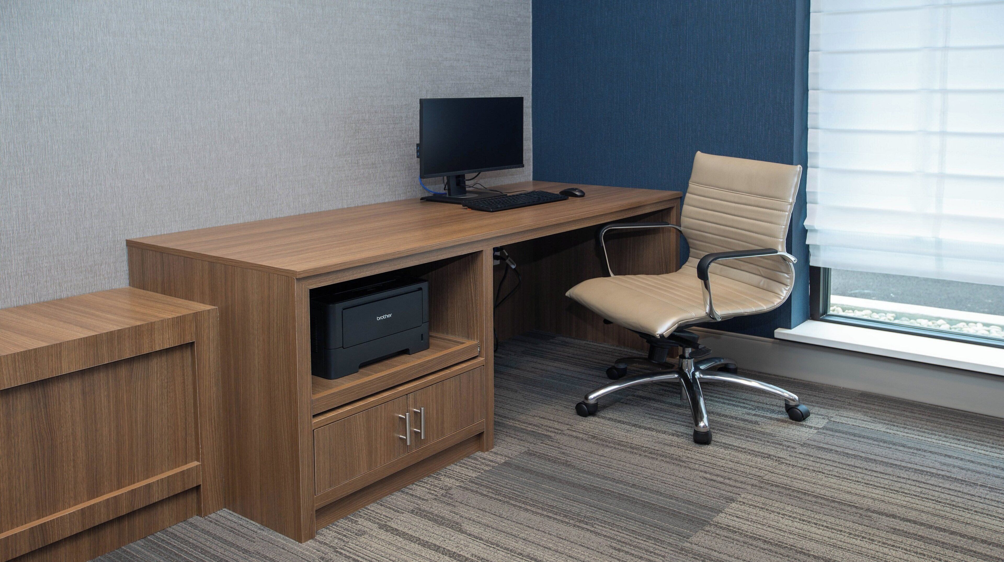 A wooden desk with a computer monitor, keyboard, and mouse, a beige office chair, and a printer in a room with a carpeted floor and a window with blinds.