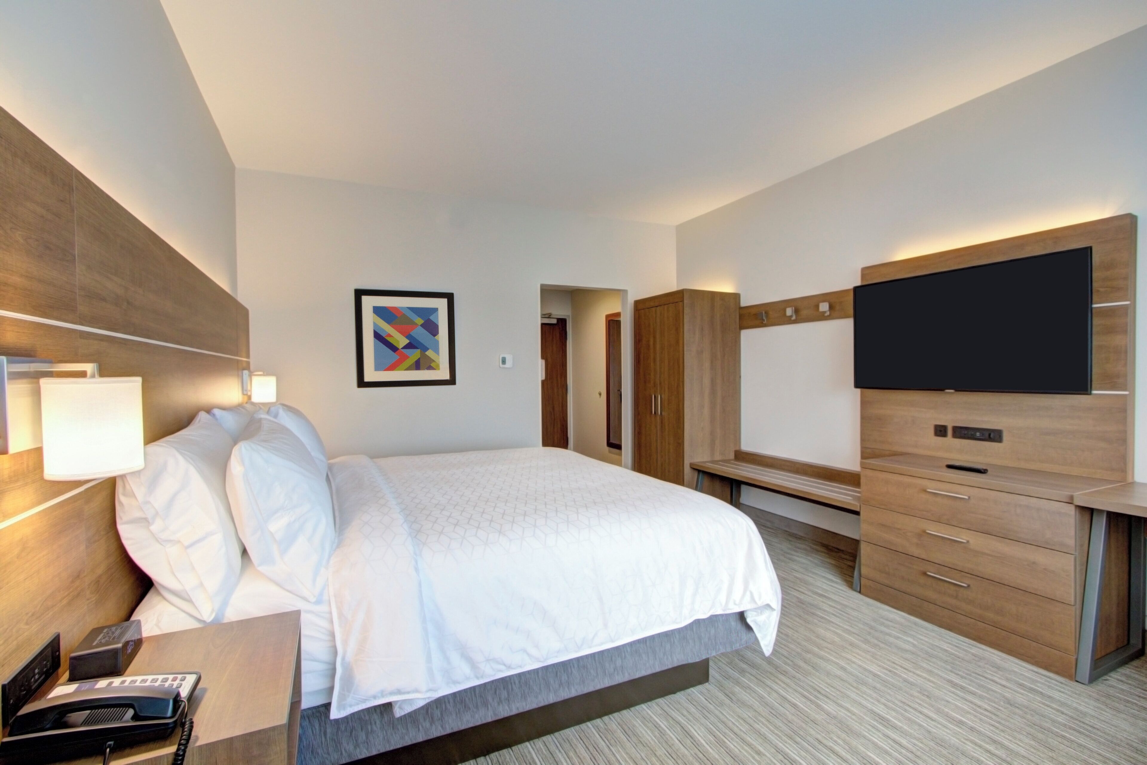 A hotel room with a bed, bedside table, lamp, TV, wardrobe, and a painting on the wall.