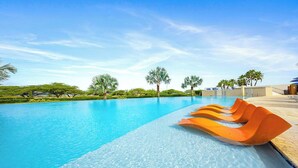 A clear swimming pool with a few orange loungers, surrounded by greenery and palm trees. - Aruba Family Friendly Oceanview PH with Hot Tub (Eagle Beach)
