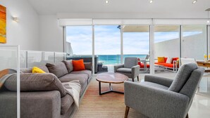 A modern living room with a large glass wall overlooking the ocean, furnished with a sofa, armchairs, and a coffee table. - Aruba Family Friendly Oceanview PH with Hot Tub (Eagle Beach)