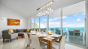 A modern dining area with a wooden table set for breakfast, large windows offering a view of the ocean, and a gray sofa with yellow cushions. - Aruba Family Friendly Oceanview PH with Hot Tub (Eagle Beach)