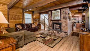 A cozy living room with a fireplace, wooden ceiling beams, a stone accent wall, and a view into the kitchen.