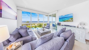 Un soggiorno moderno con un'ampia finestra che si affaccia su una vista mare panoramica, divani rivestiti in grigio, un tavolino da caffè e un mobile contenitore bianco con piano in vetro.