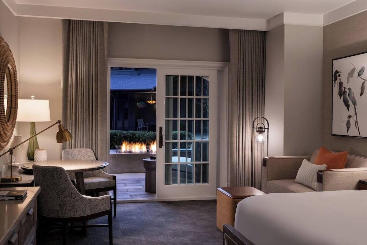 A hotel room with a bed, a desk, a chair, a table, a lamp, a painting, and a view of the outdoors.