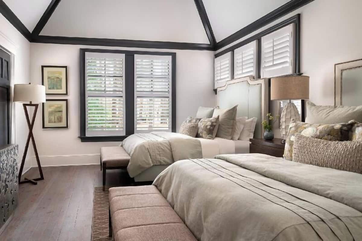 A bedroom with two beds, a bench, a floor lamp, and a window with shutters.