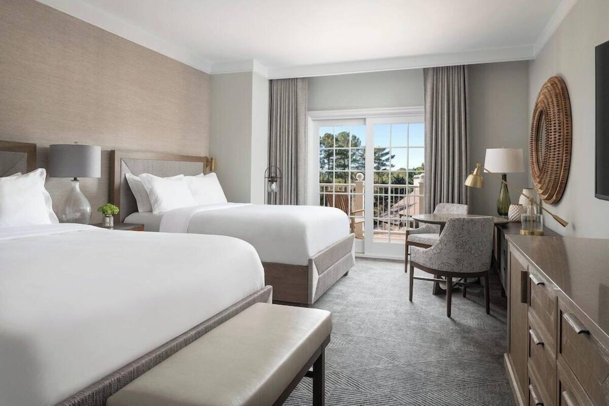 A hotel room with two beds, a desk, a chair, a TV, and a balcony with a view.