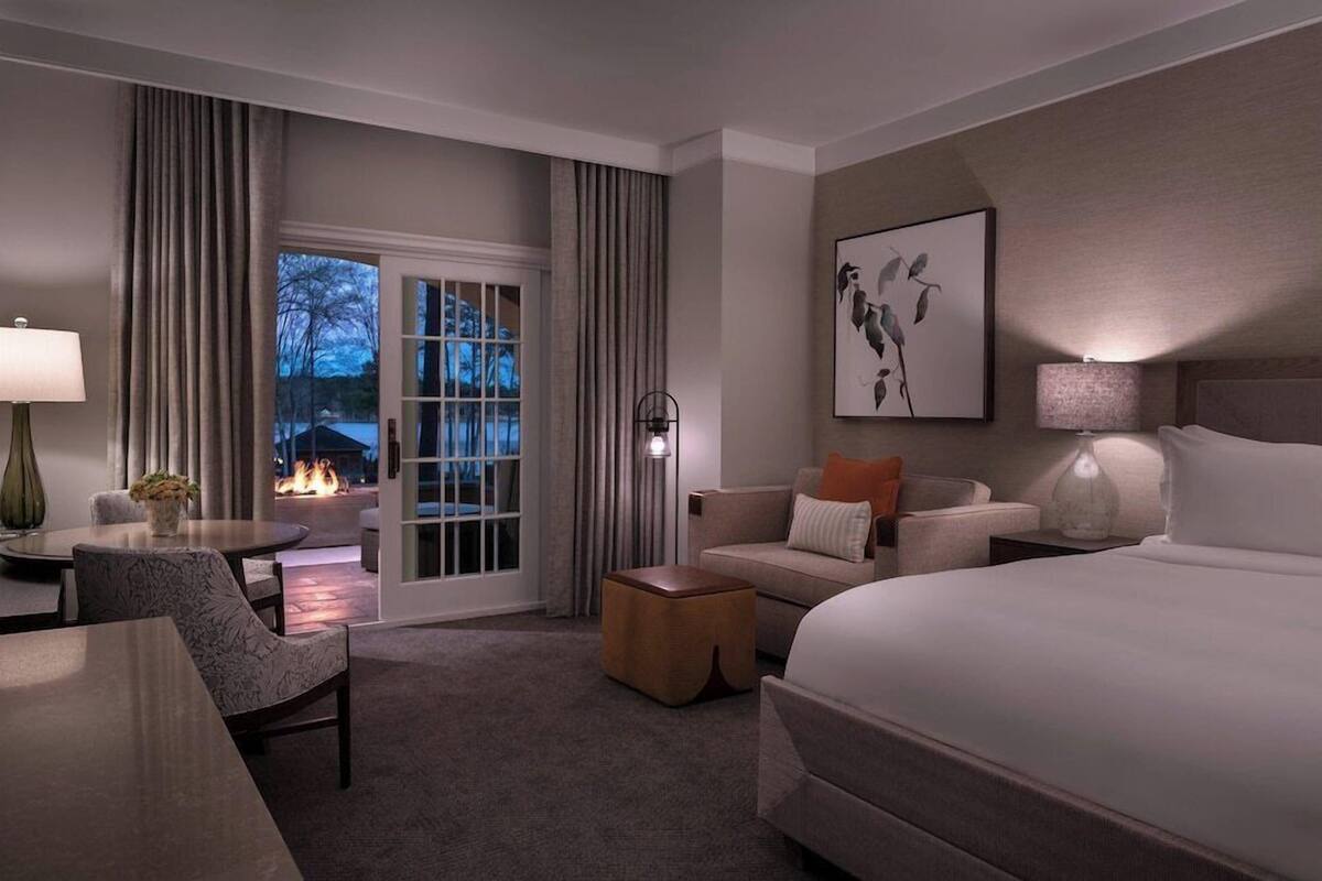 A hotel room with a bed, a sofa, a chair, a small table, a lamp, a painting, and a view of the outdoors.