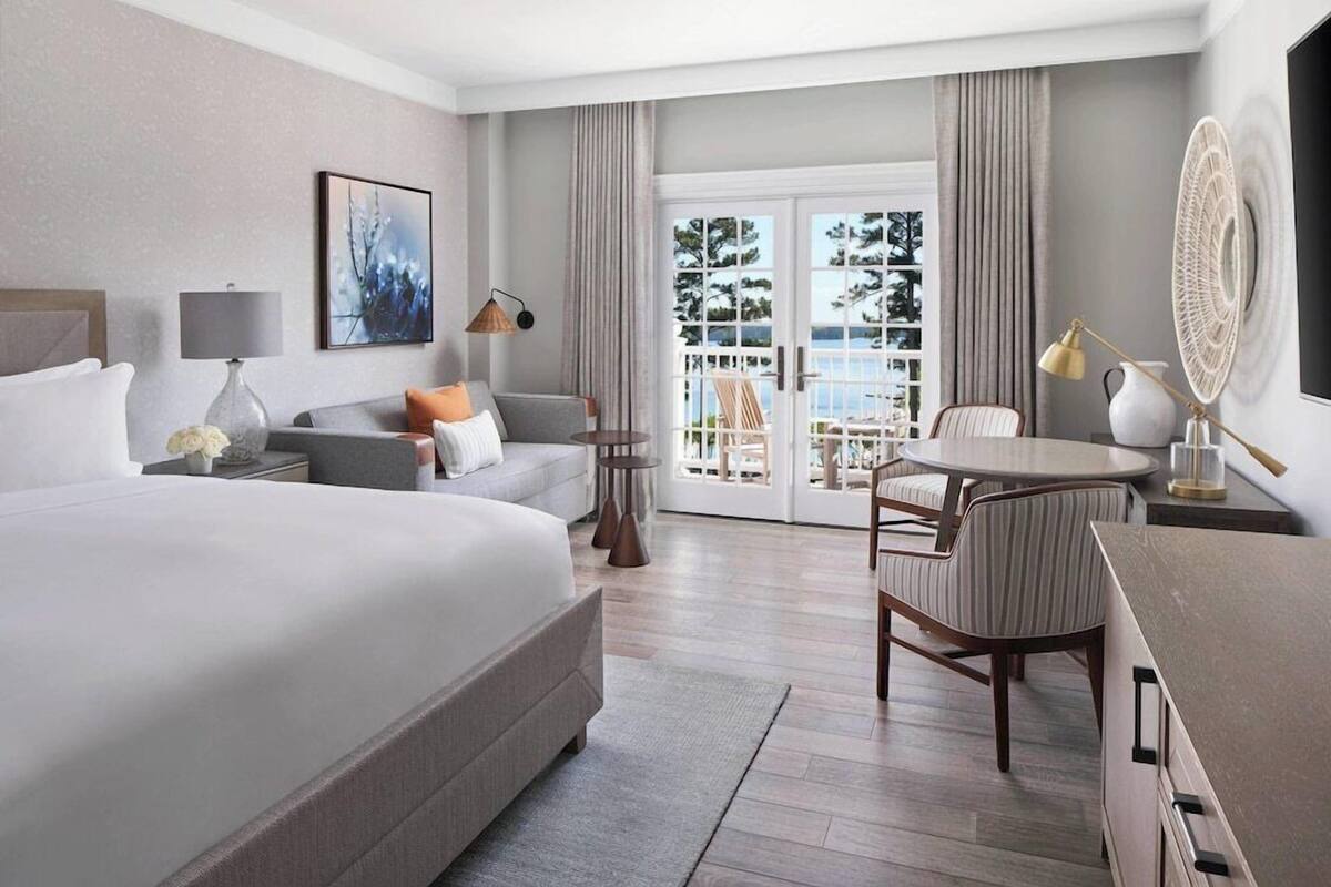 A hotel room with a large bed, a seating area with a table, a TV, and a view through a glass door.