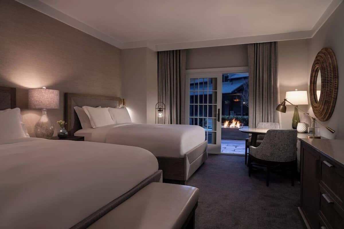 A hotel room with two beds, a desk, a chair, a fireplace, and a view of the outside.