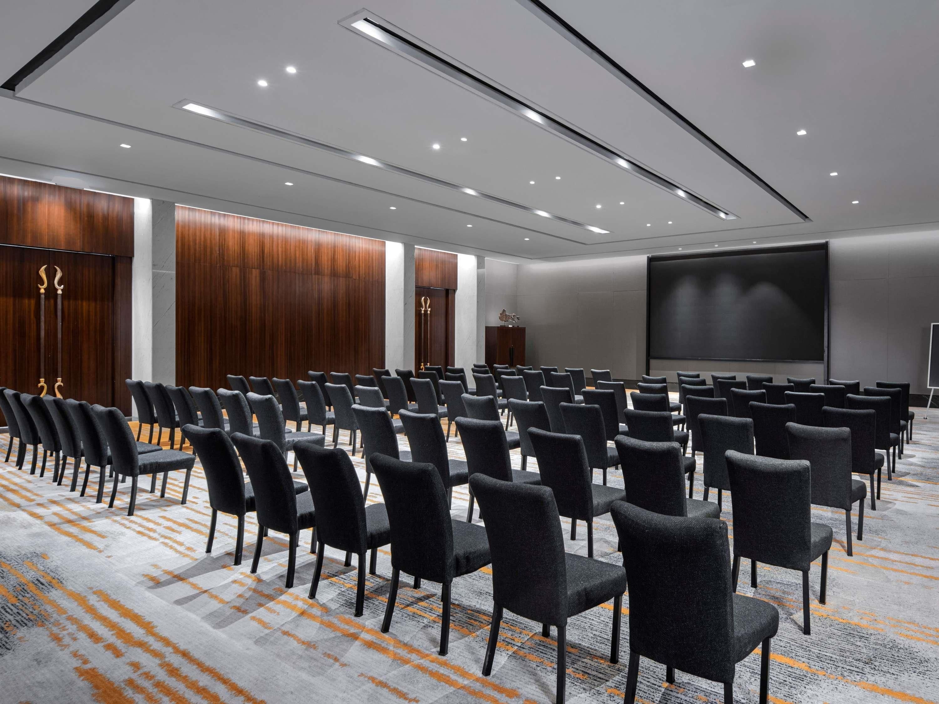 Un salón de conferencias con filas de sillas, una pantalla grande y suelo alfombrado.