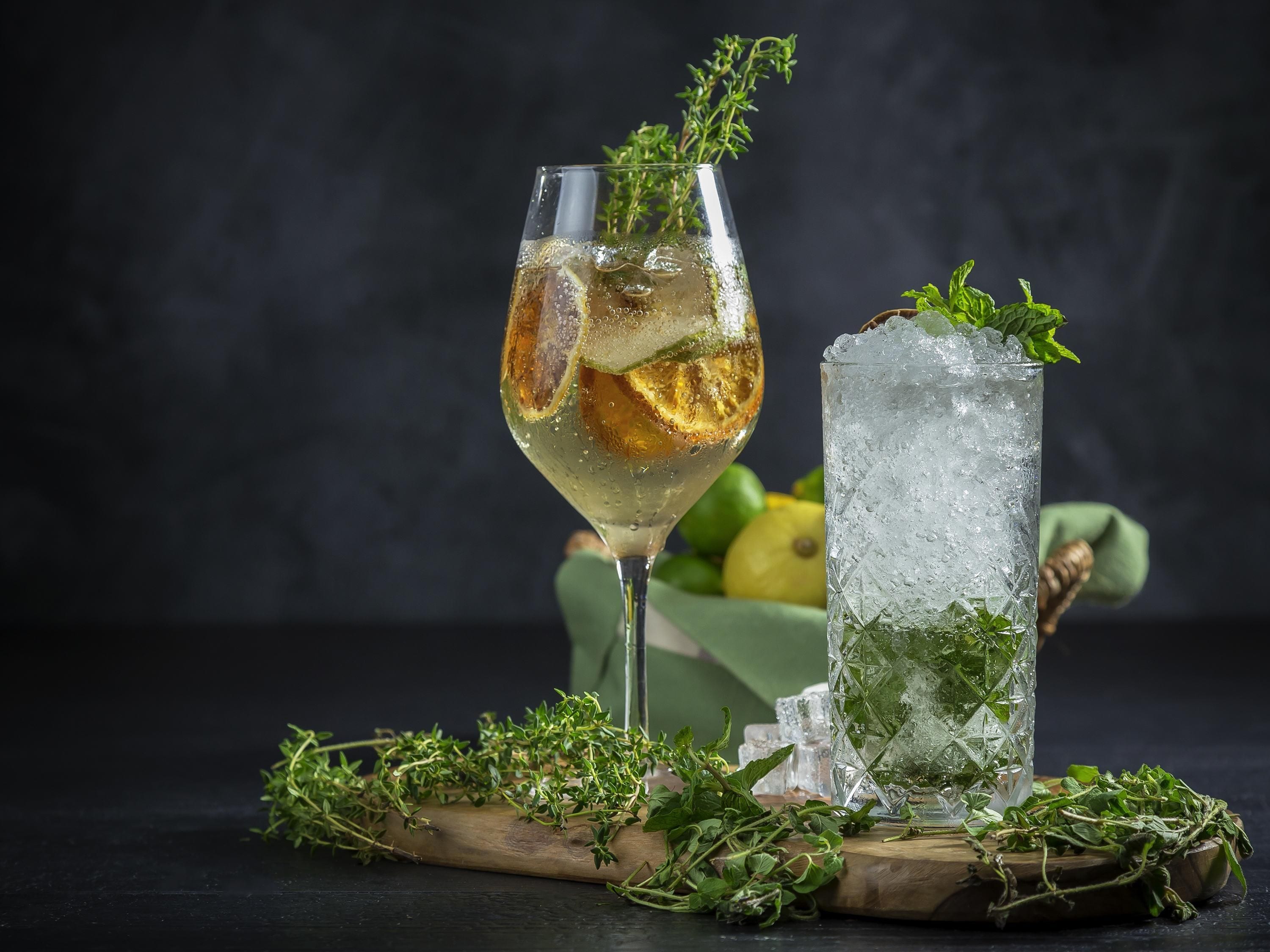 Two glasses of a beverage with ice and herbs, a wooden board with more herbs, and a bowl of lemons.
