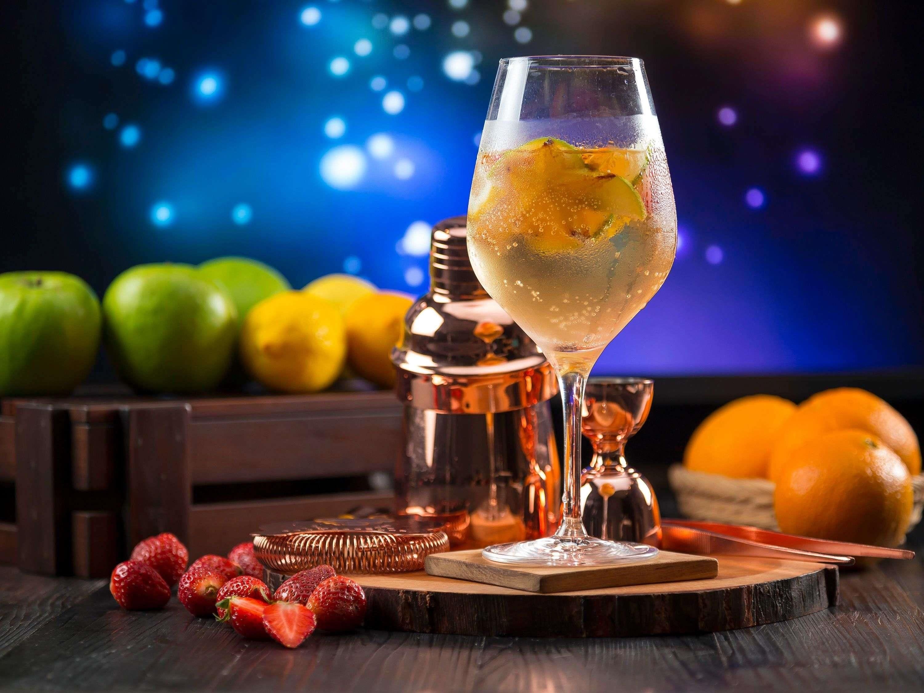 A cocktail glass with a lime slice on a wooden coaster, surrounded by fruits and a copper shaker.