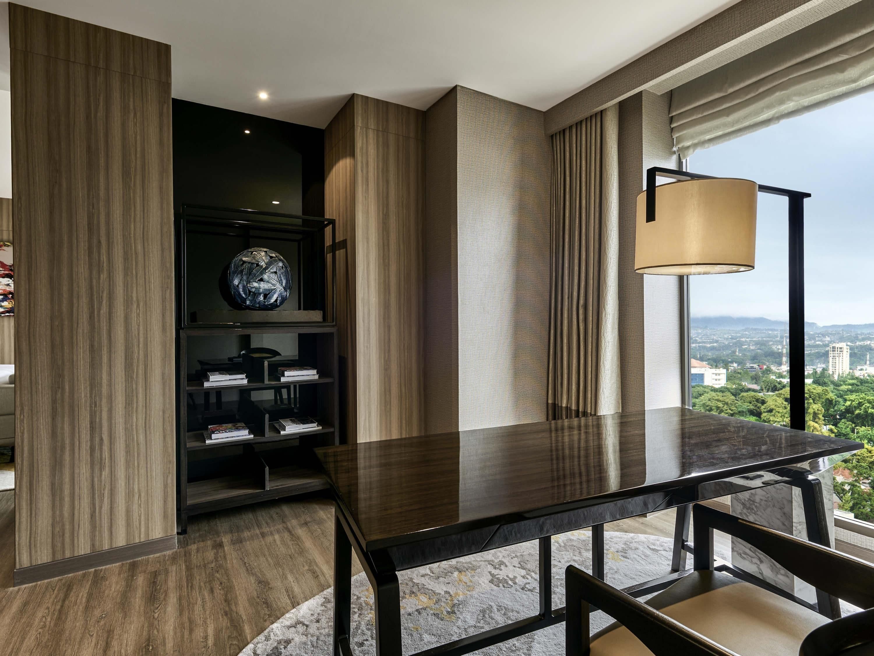 A modern hotel room with a dark wood floor, a marble-topped table, a bookshelf, and a large window offering a city view.