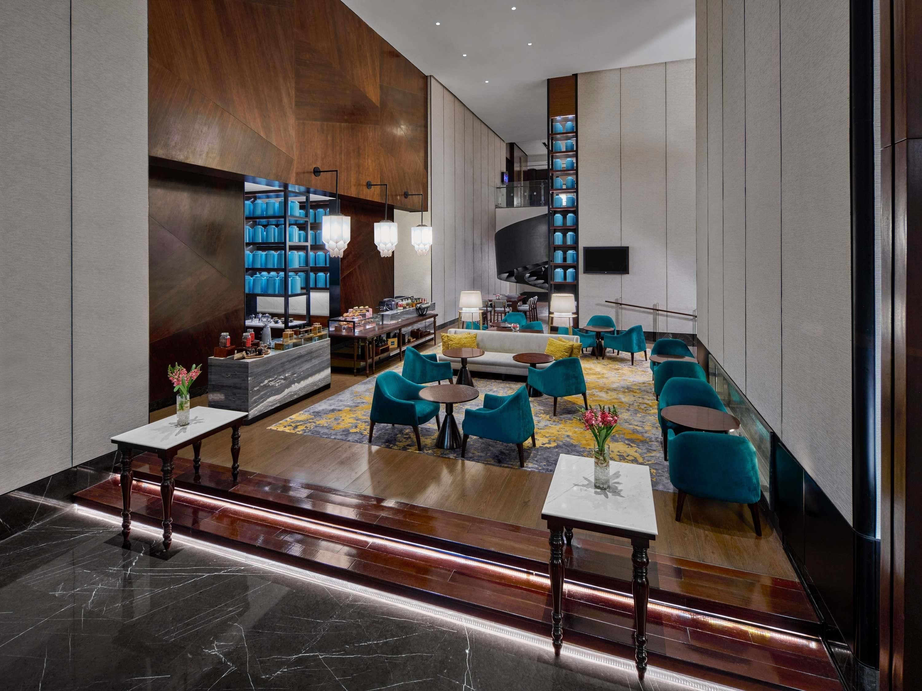 A modern lobby with a marble floor, wooden paneling, a seating area with teal chairs, a bar with blue storage, and a flat-screen TV mounted on the wall.