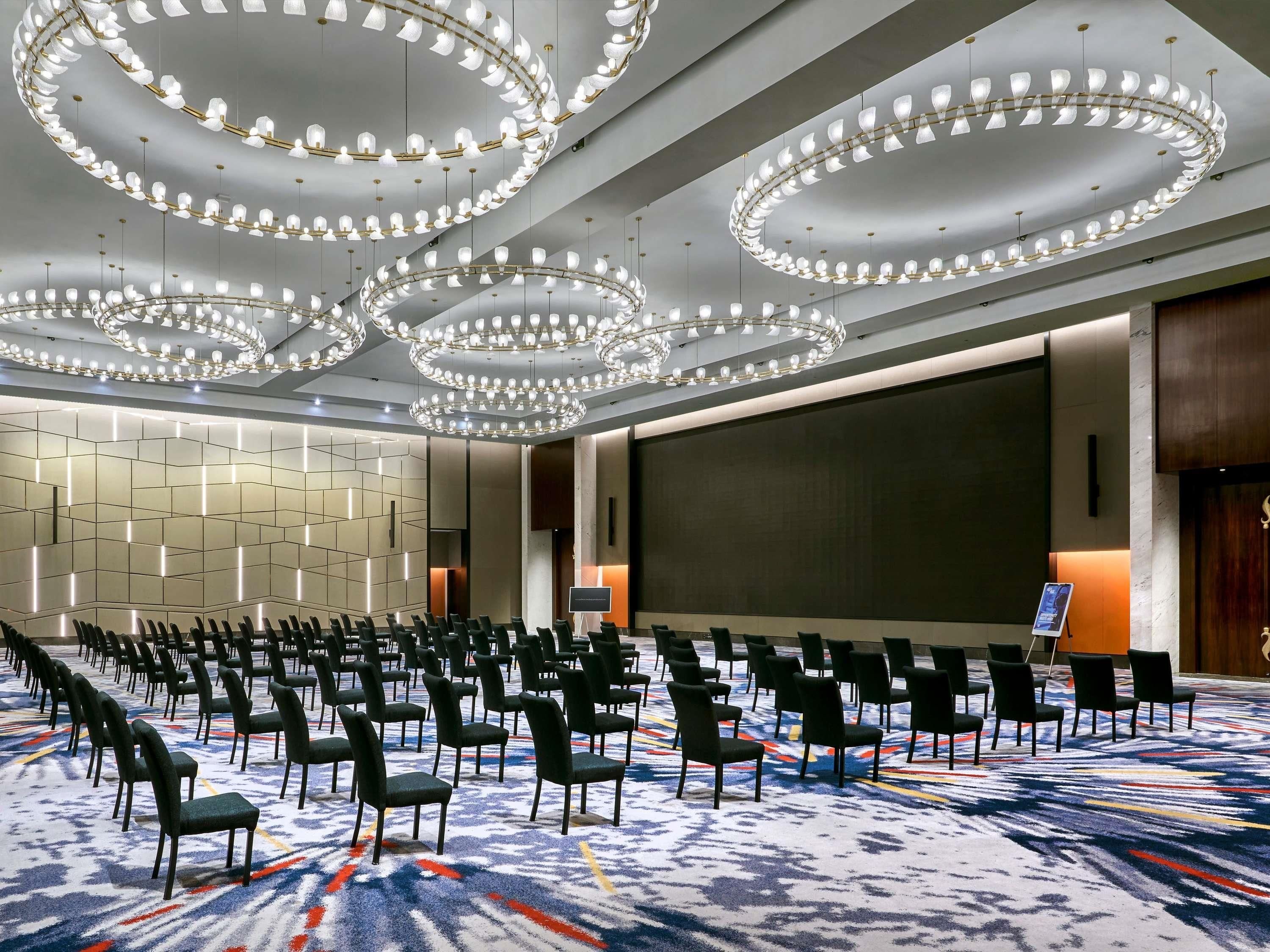 A conference room with rows of chairs, a large screen, and multiple circular chandeliers.