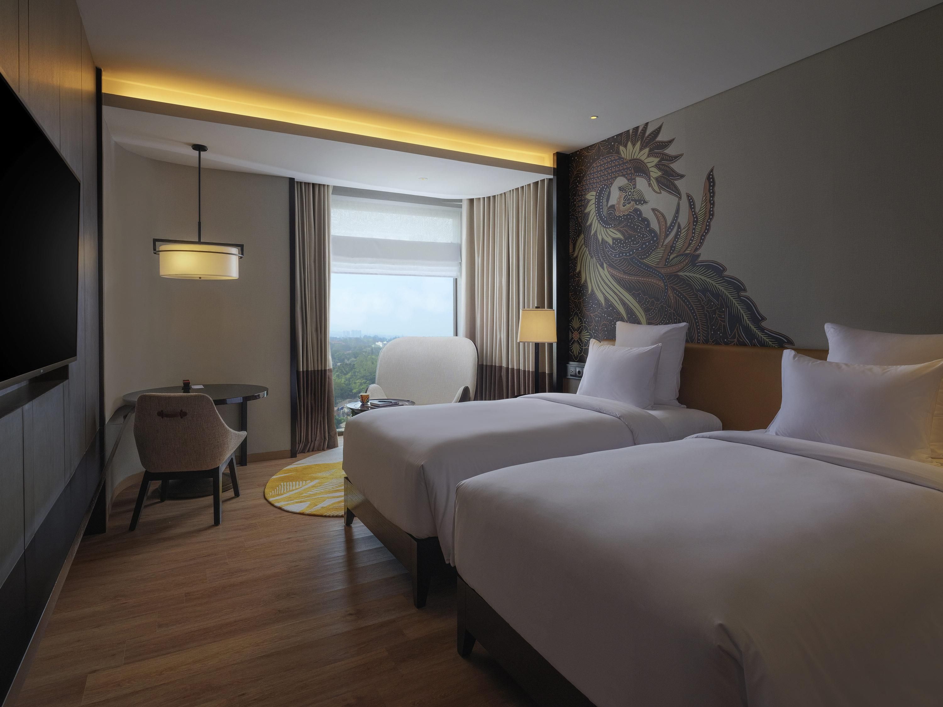 A hotel room with two beds, a desk, a chair, a TV, and a large window with curtains.