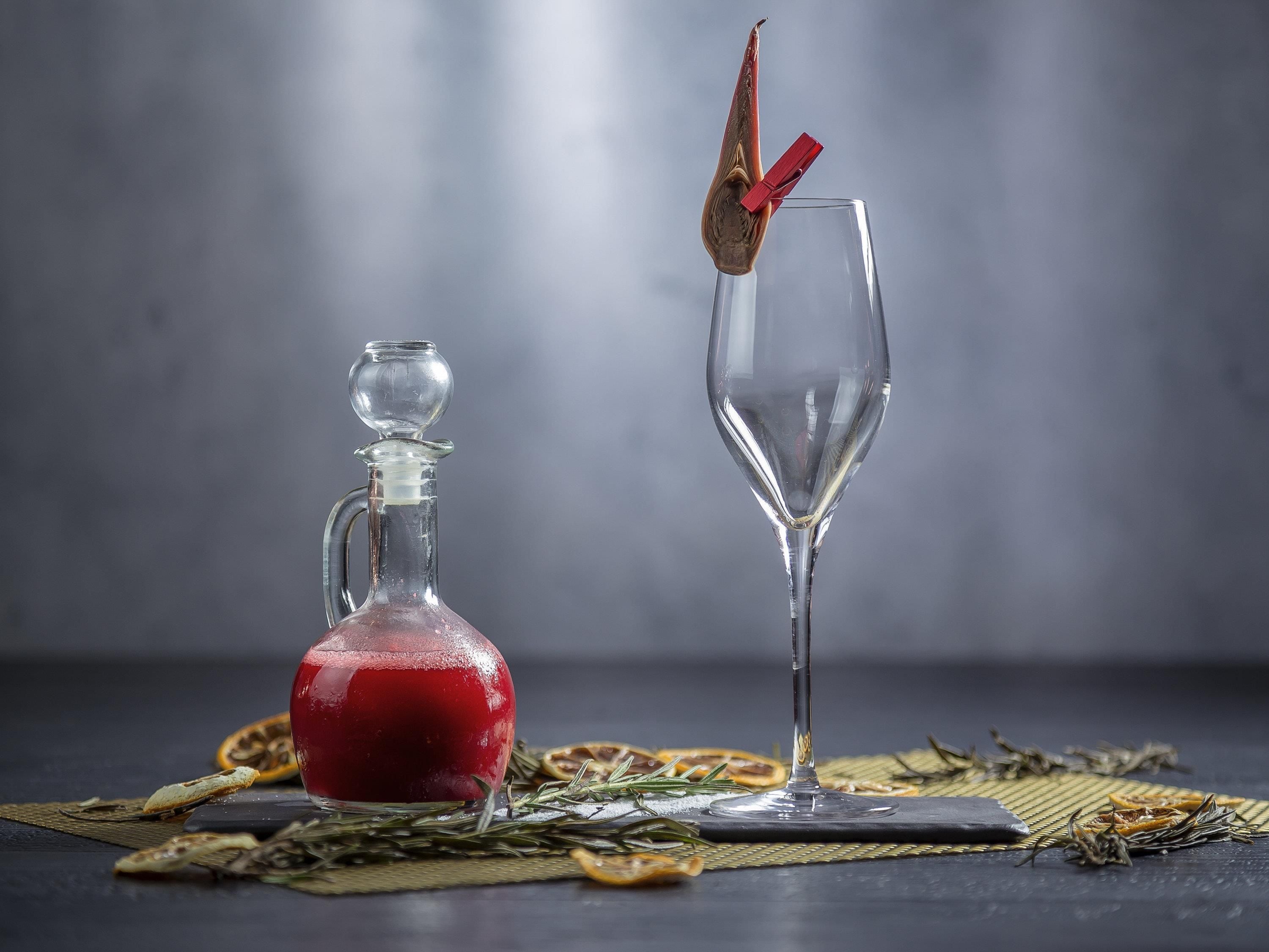 A glass decanter and a wine glass with a red clothespin on a table with dried herbs.