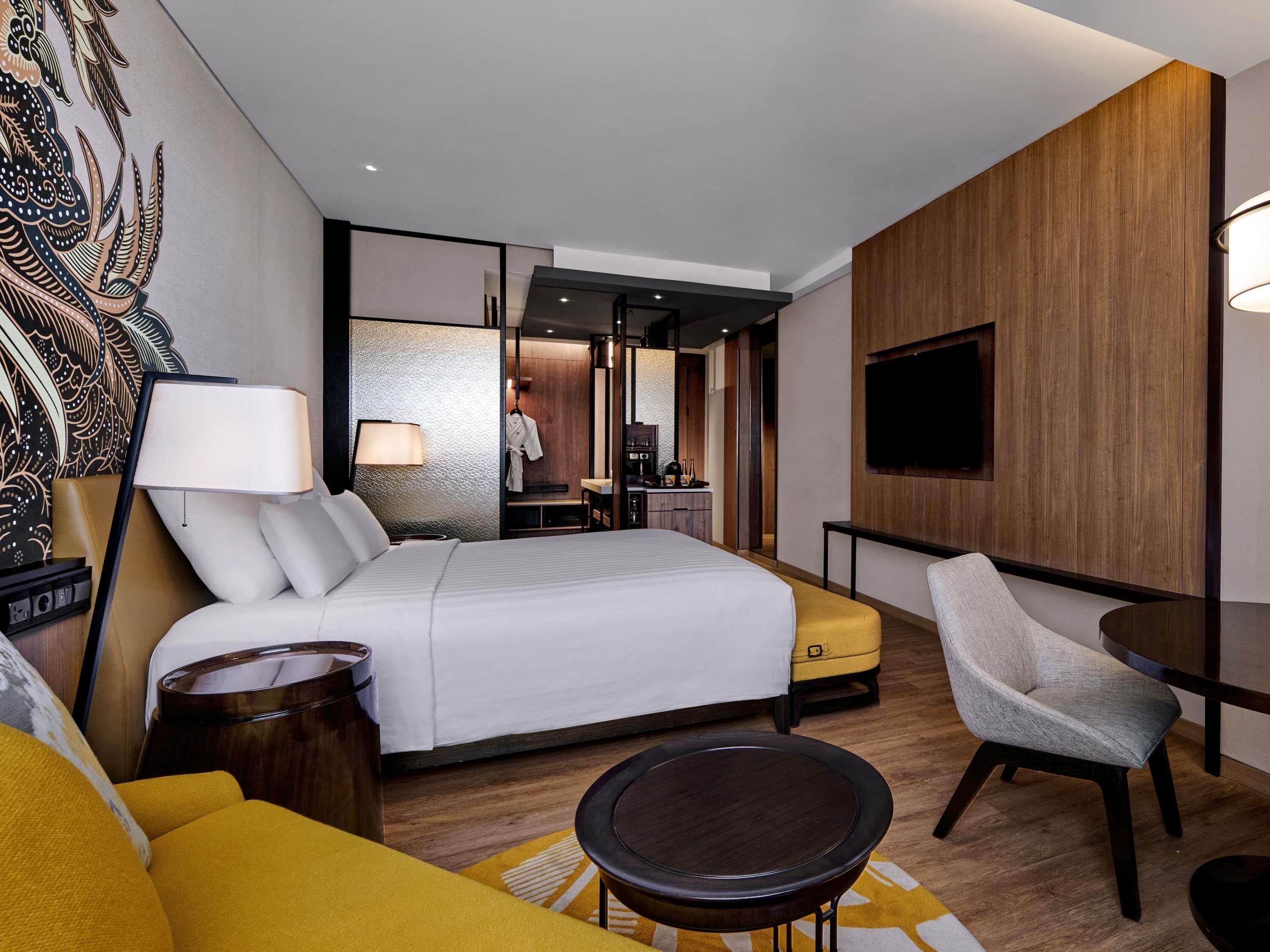 Una habitación de hotel moderna con una cama grande, un sofá, una silla, una mesita y un televisor montado en la pared.