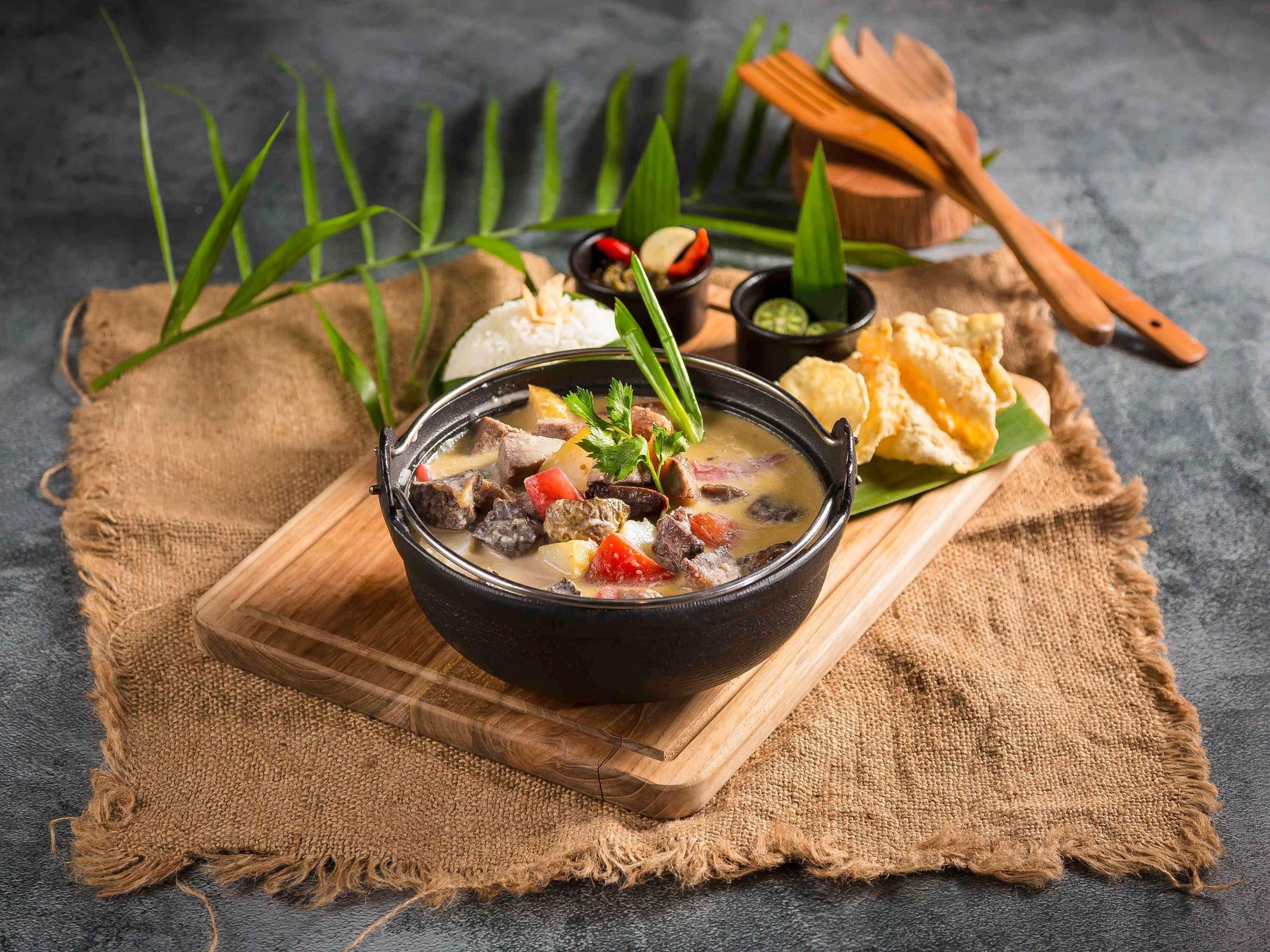 A bowl of stew with vegetables and meat, accompanied by a side of bread and dipping sauces on a wooden board.