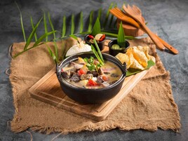 Un bol de ragoût de légumes et de viande, servi avec du pain et des sauces pour tremper, le tout présenté sur une planche en bois.
