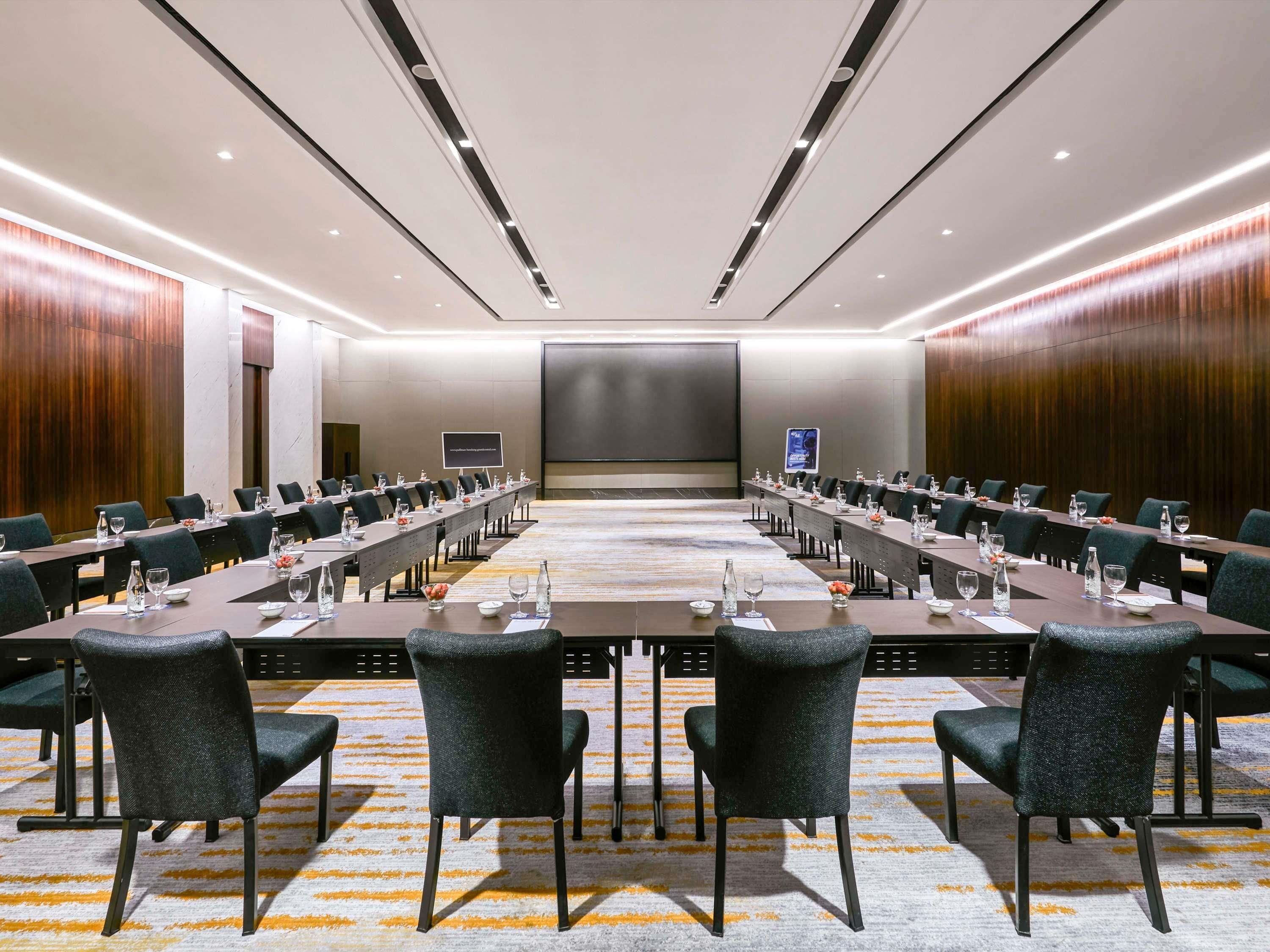 Een vergaderzaal met lange tafels, stoelen en een groot scherm aan de voorkant.