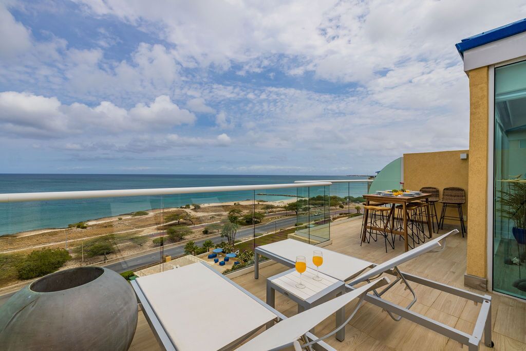 Un balcón con vistas a la playa y al océano, mobiliario exterior y un ventanal grande.