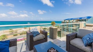 A balcony with wicker furniture, a small tree planter, and a view of the beach and ocean.