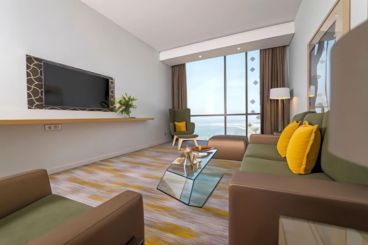 A modern living room with a flat-screen TV, comfortable seating, a coffee table, and a large window offering a view.