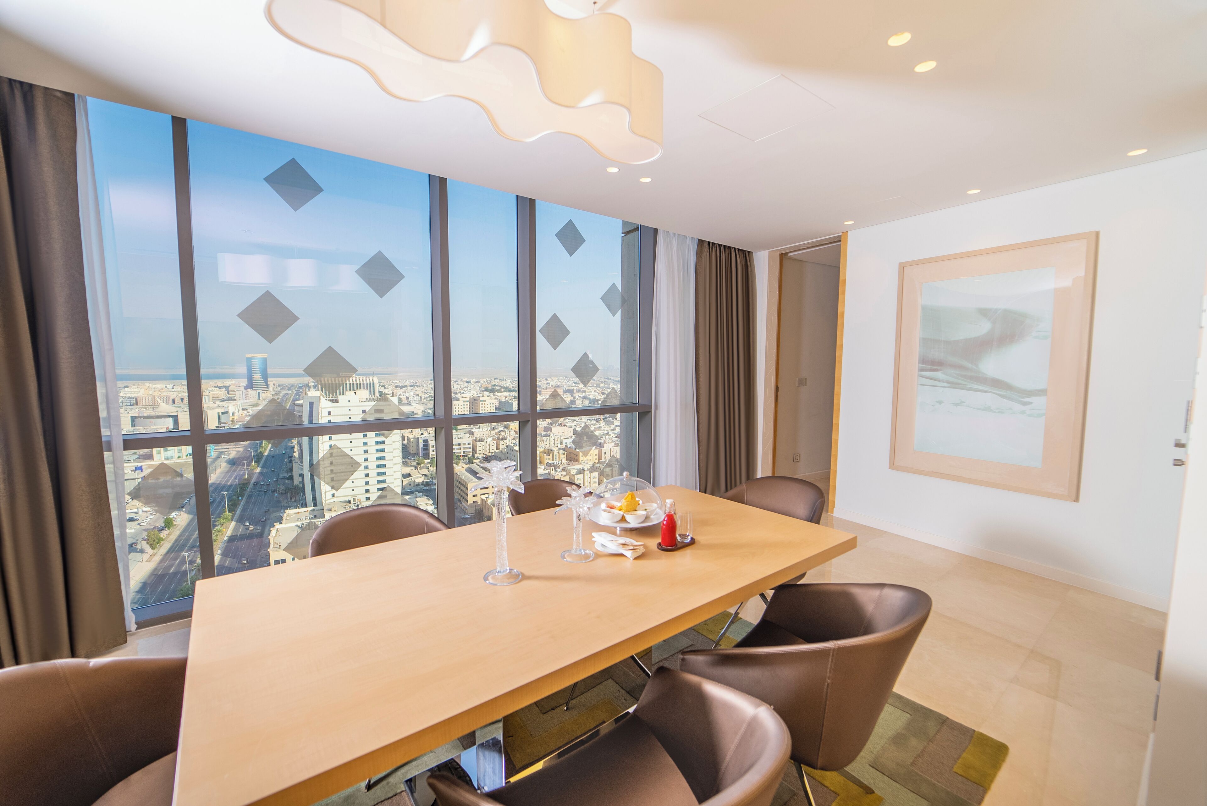 A modern hotel room with a large window offering a city view, a wooden dining table, and brown leather chairs.