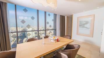 A modern hotel room with a large window offering a city view, a wooden dining table, and brown leather chairs.