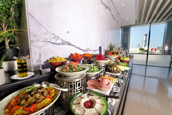 A buffet with various dishes, a marble wall, and a view of a cityscape through large windows. - Rotana Corniche (Dammam)