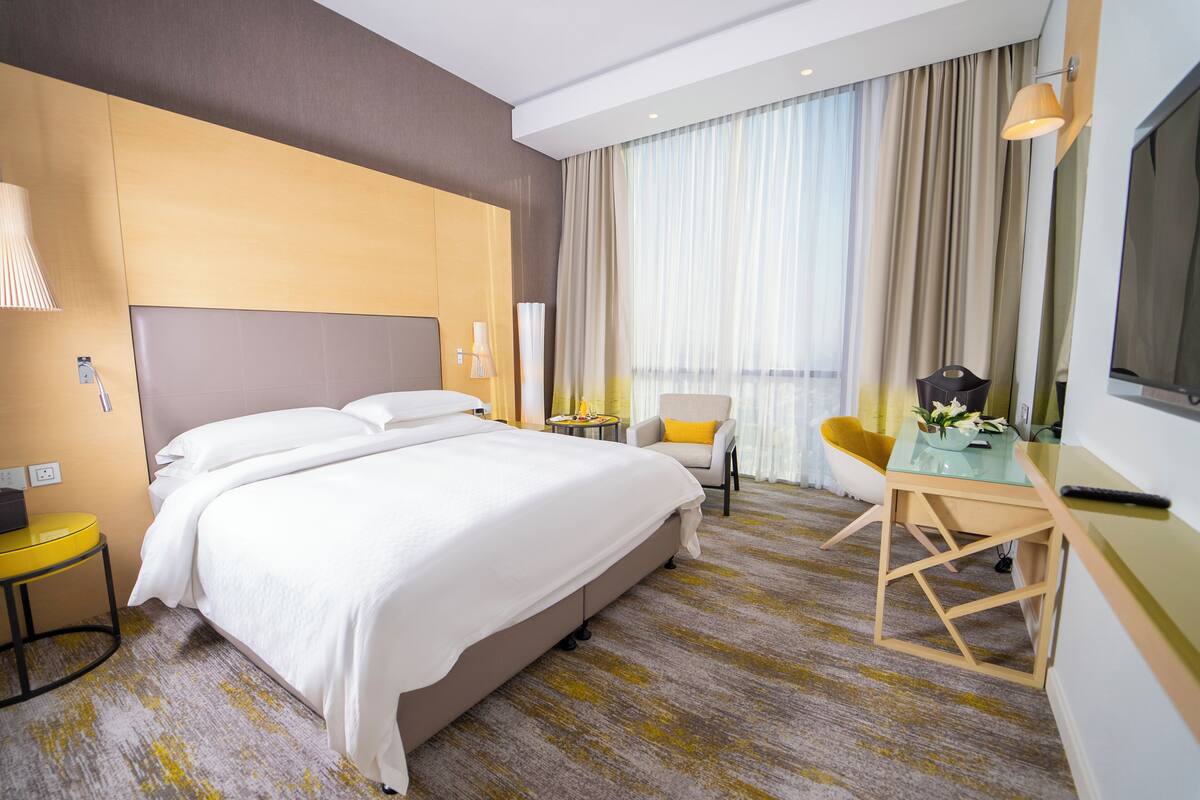 A hotel room with a large bed, a desk, a chair, a lamp, and a TV.