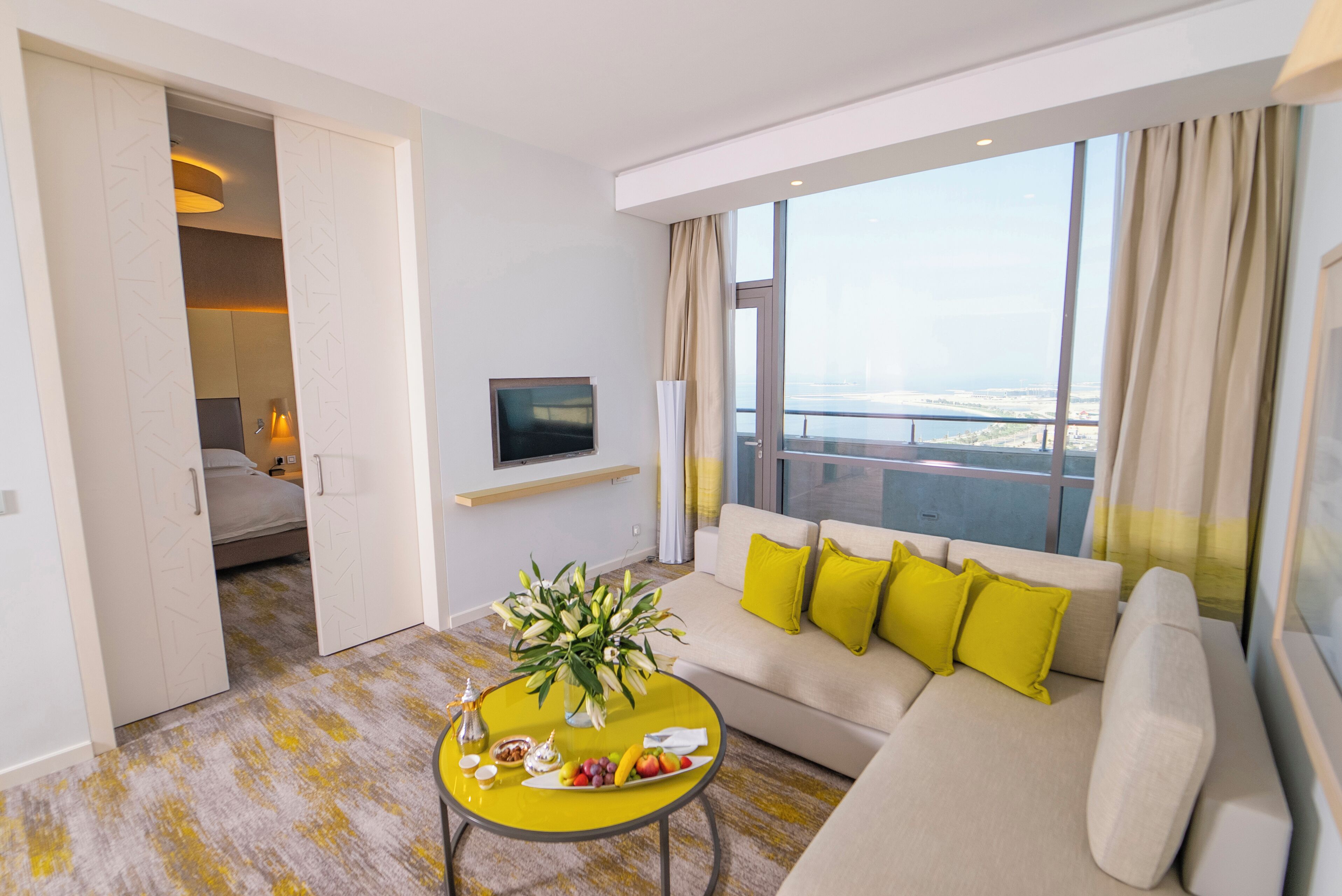 Une chambre d’hôtel moderne avec un canapé, une table basse agrémentée de fruits et d’un vase, une télévision et un balcon offrant une vue agréable.