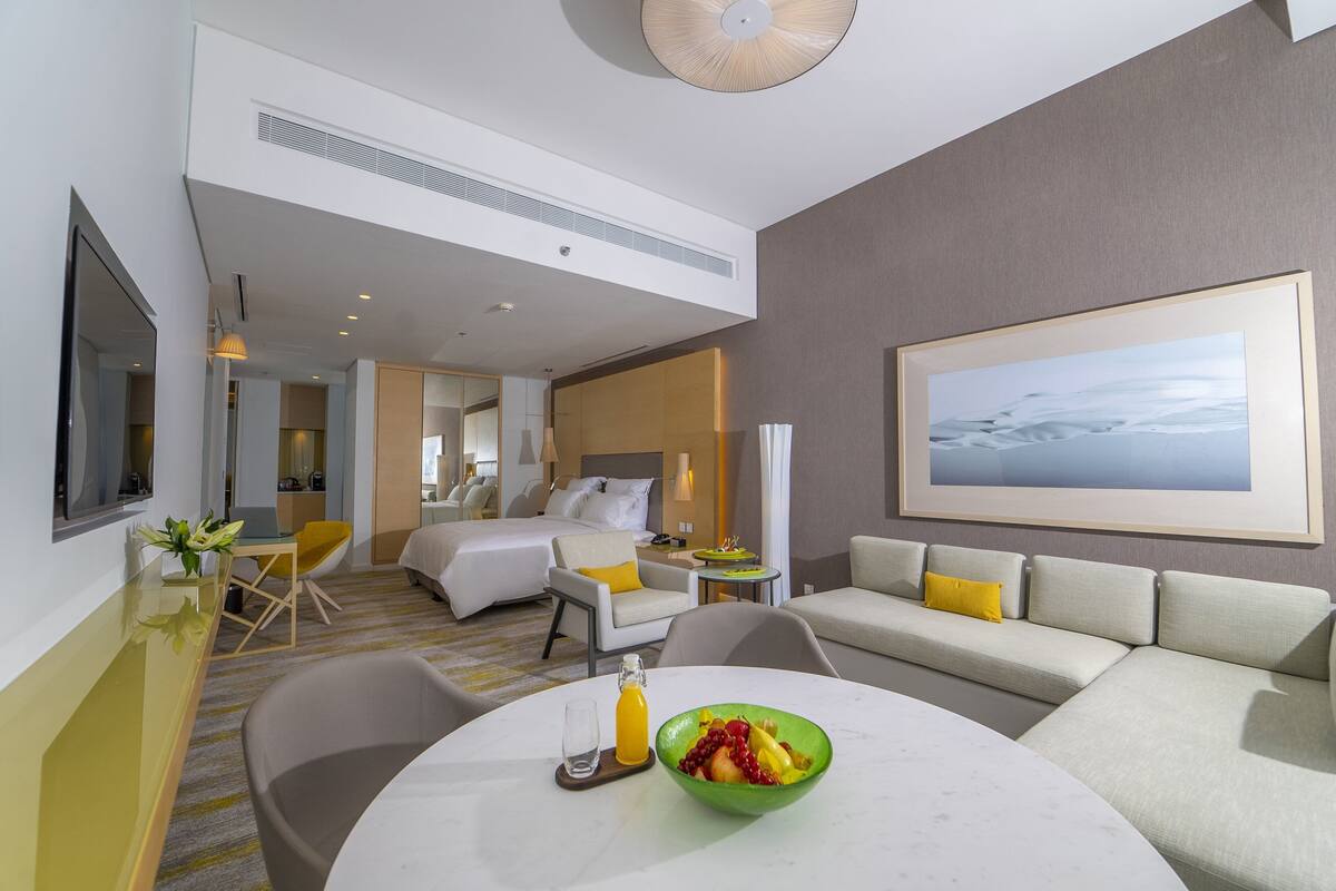 A modern hotel room with a bed, a sofa, a dining table with fruit, and a painting on the wall.
