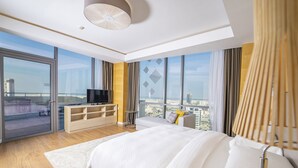 Una habitación de hotel moderna con una cama grande, un televisor sobre un mueble de madera, un balcón con vistas a la ciudad y un ventanal con cortinas.