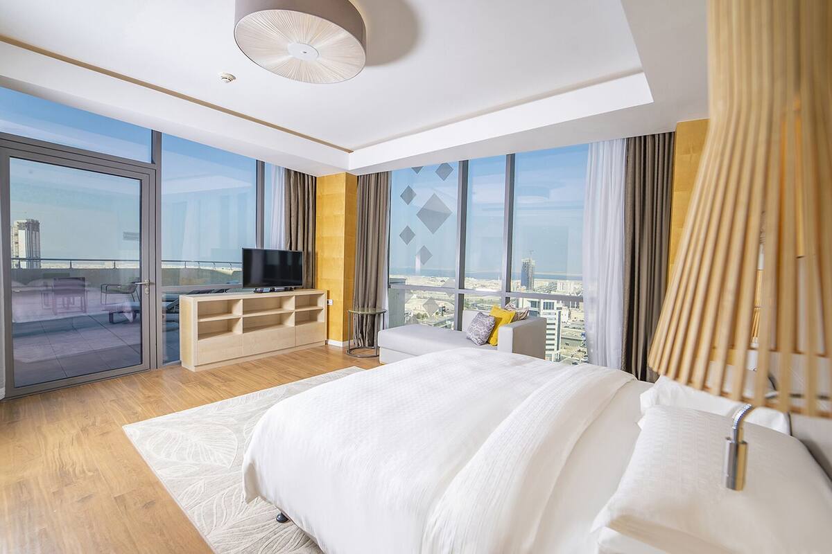 A modern hotel room with a large bed, a TV on a wooden console, a balcony with a city view, and a large window with curtains.