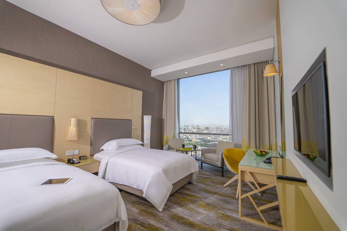 A hotel room with two beds, a desk with a chair, a TV, and a view of the city.