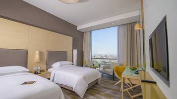 Quarto de hotel com duas camas, uma escrivaninha com cadeira, televisão e vista para a cidade.