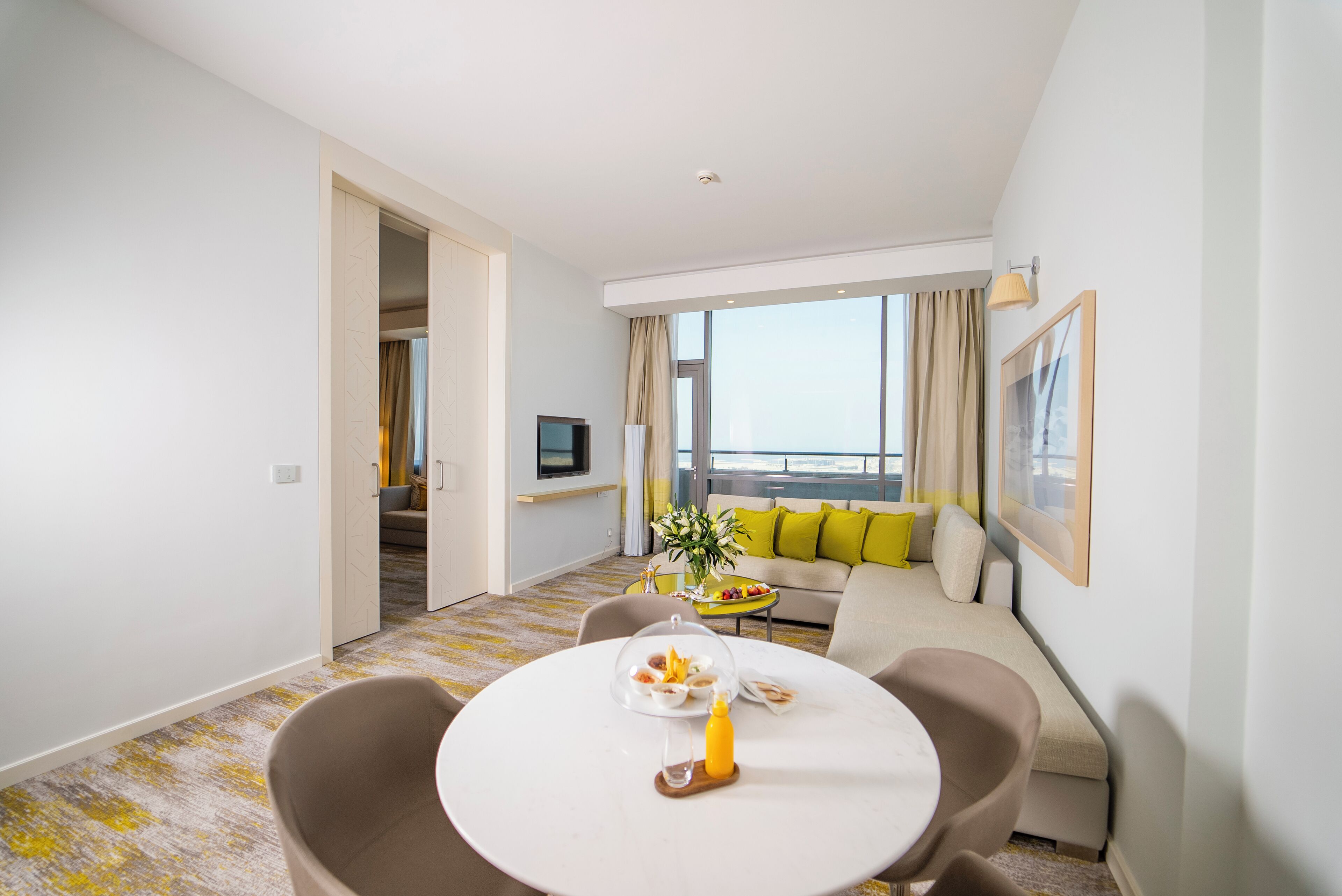A modern hotel room with a dining area, a sofa, a TV, and a balcony with a view.