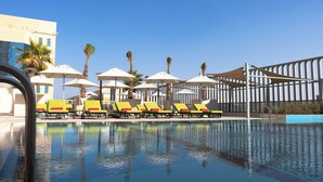 Una piscina con sillas de descanso y sombrillas, rodeada de palmeras y un edificio moderno.